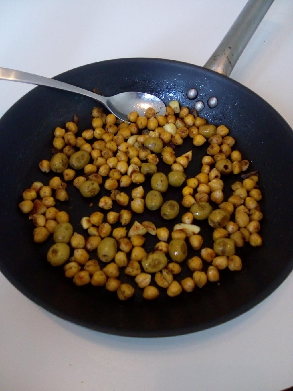 Picture of chickpeas, olives and chopped garlic fried in a pan, with a spoon